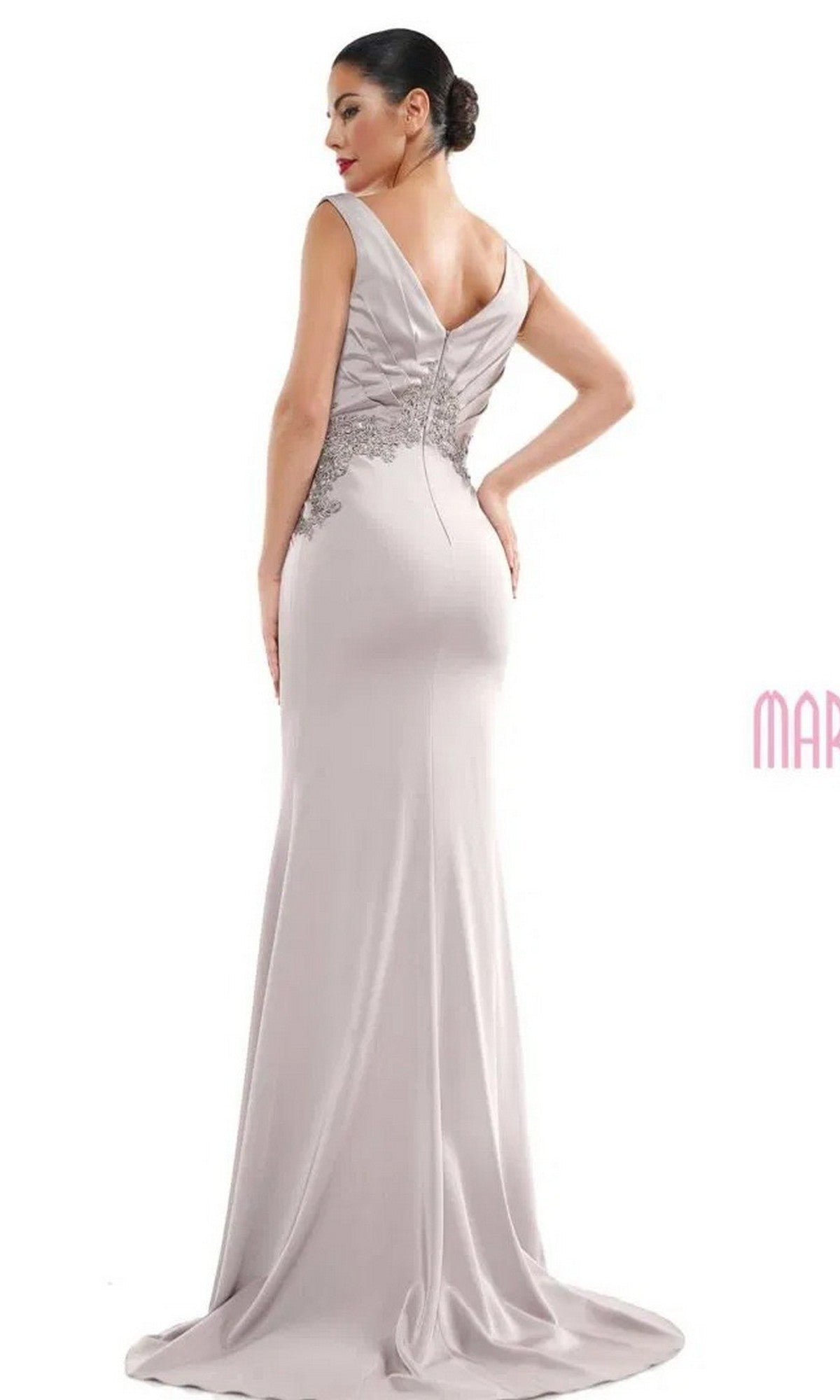 A woman wears the Marsoni MV1054 long formal dress in light gray, featuring a sleeveless design, V-back, embroidered floral lace bodice, and a trumpet silhouette. Her hair is styled in a low bun as she stands against a plain background.
